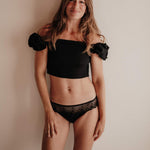 Frau trägt eine schwarze Perioden-Panty im hüftigen Hipster-Style mit extra hoher Sauglage für Frontbleeder. Aus atmungsaktivem TENCEL™ Lyocell, hautfreundlich, mit recycelter Spitze am Bund, fair in Portugal gefertigt.